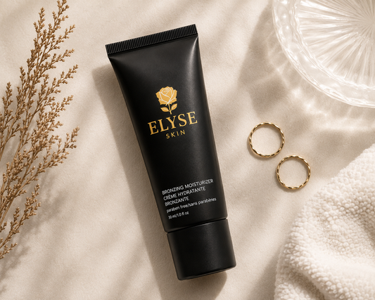 A black squeeze tube of Elyse Skin Bronzing Moisturizer lies on a soft cream fabric, styled with dried botanicals, two gold rings, and a clear textured glass dish.