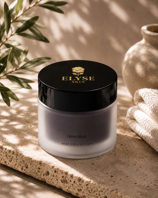 A jar of ELYSE SKIN Glow Mask sits on a textured stone slab with olive branches and a neutral ceramic vase in the background, creating a warm, natural aesthetic.
