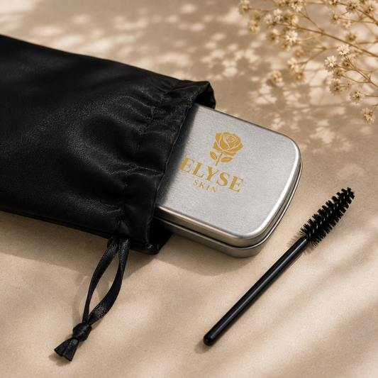 A silver metal cosmetic case with a gold rose logo and the brand name 'ELYSE SKIN' partially pulled from a black satin drawstring pouch, with a black spoolie brush placed beside it on a beige fabric surface with soft leaf shadows.