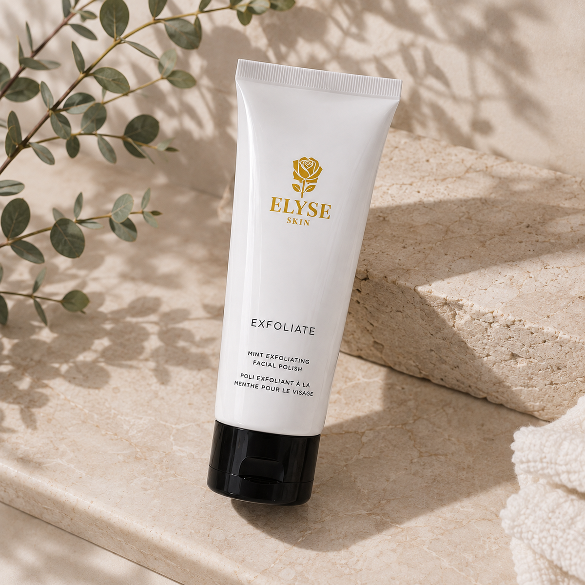 A white squeeze tube of ELYSE SKIN Exfoliate facial polish with a black cap, styled on a beige stone surface with eucalyptus leaves and soft shadows.