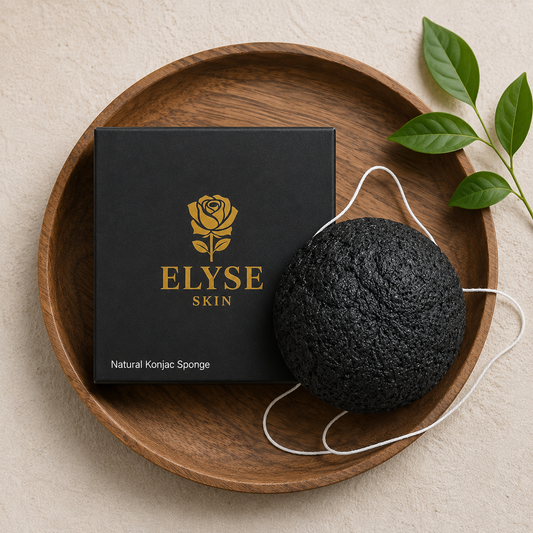 A black textured natural konjac sponge with a white elastic loop, displayed next to its black and gold ELYSE SKIN packaging box on a round wooden tray.