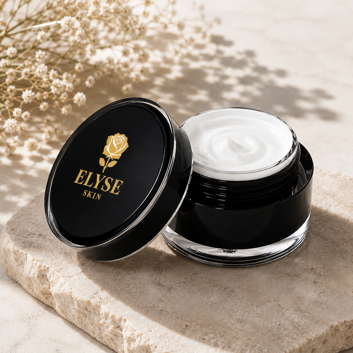 An open black cosmetic jar with white cream inside, next to its matching black lid featuring a gold rose logo and the brand name 'ELYSE SKIN', styled on a beige stone slab with dried flowers in the background.