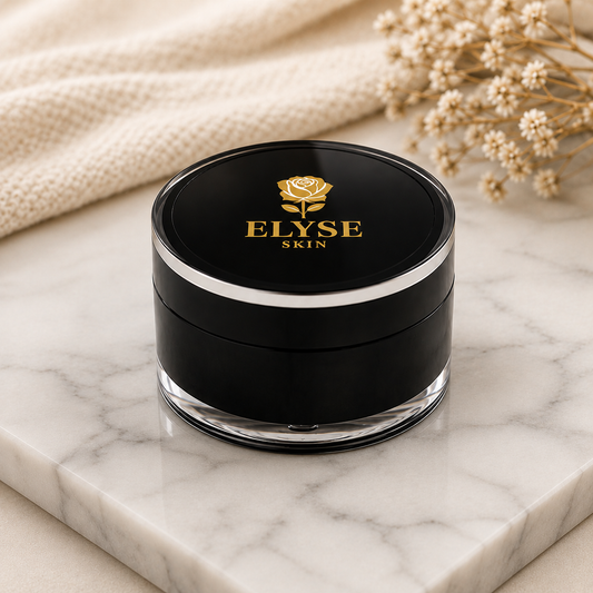 A black cosmetic jar with a clear base and a gold rose logo reading ELYSE SKIN on the lid, placed on a white marble surface with a beige knit fabric and dried flowers in the background.