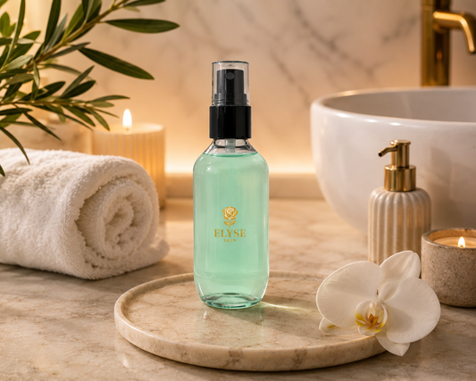 A clear bottle of ELYSE PARIS body mist filled with light aqua liquid, standing on a round marble tray in a spa-like bathroom setting with a rolled towel, candle, orchid flower, and sink in the background.