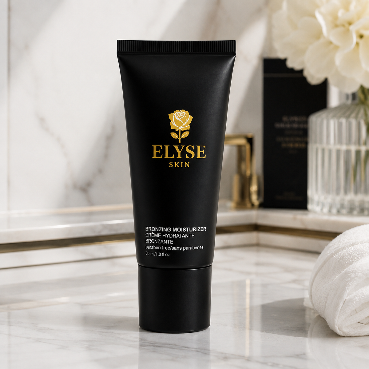 A black squeeze tube of ELYSE SKIN Broadening Moisturizer stands on a white marble surface with a softly blurred bathroom-like setting behind it, including white flowers, a glass vase, and a rolled towel.