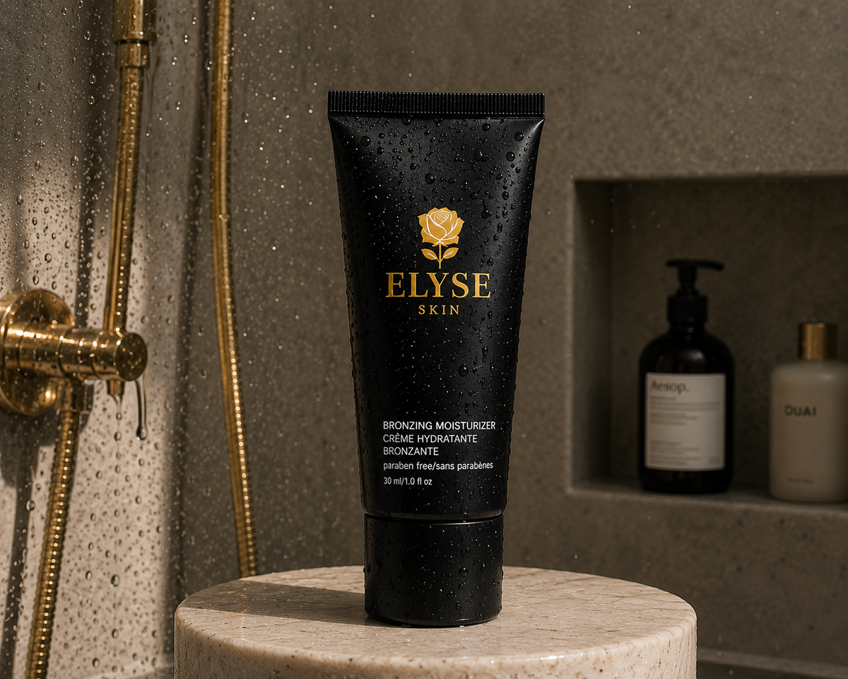A black squeeze tube of ELYSE SKIN Bronzing Moisturizer stands on a round stone pedestal in a bathroom setting. The tube has gold lettering and a gold rose logo, with water droplets on its surface.