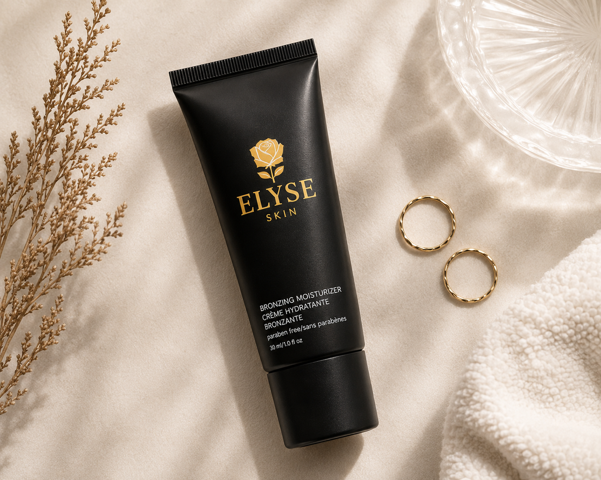A black squeeze tube of Elyse Skin Bronzing Moisturizer lies on a soft cream fabric, styled with dried botanicals, two gold rings, and a clear textured glass dish.