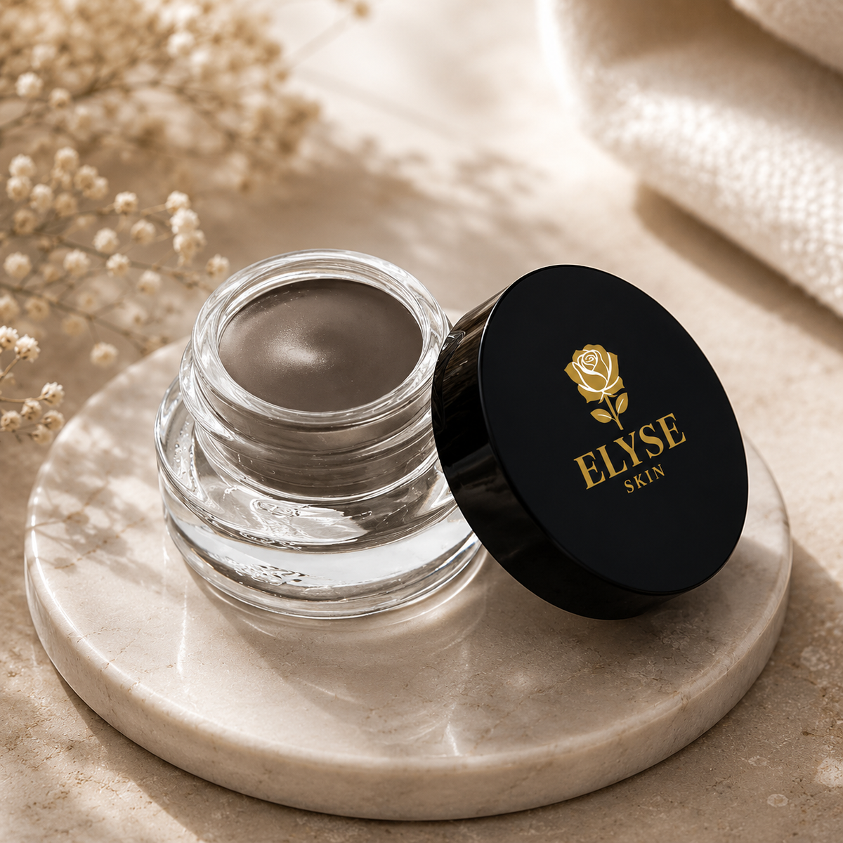 An open clear cosmetic jar with dark brown product inside, next to a black lid printed with a gold rose and the brand name ELYSE SKIN, set on a round stone tray with dried flowers and soft neutral fabric in the background.