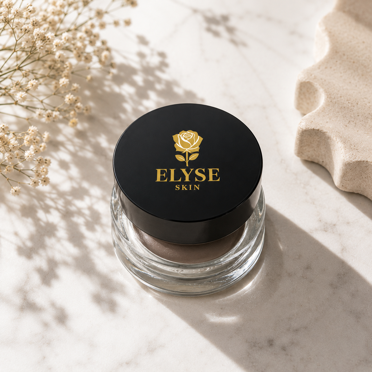 A clear cosmetic jar with a black lid featuring a gold rose logo and the text 'ELYSE SKIN', placed on a light marble surface with dried flowers and soft shadows.