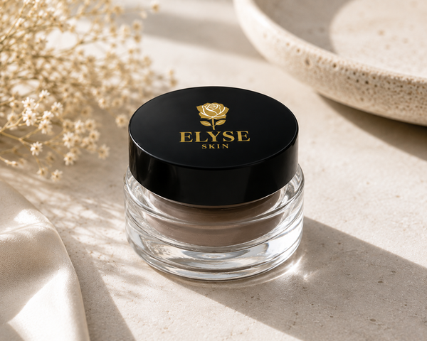 A clear cosmetic jar with a black lid and gold 'ELYSE SKIN' logo, styled on a light stone surface with dried flowers and soft shadows.