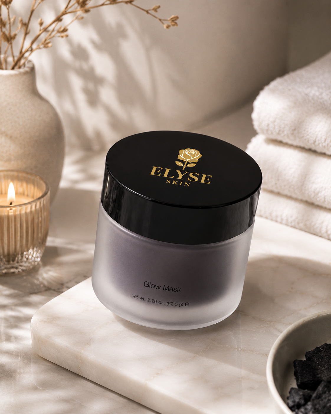 A jar of ELYSE SKIN Glow Mask sits on a white marble tray in a spa-like setting with folded white towels, a lit candle, and dried branches in the background.