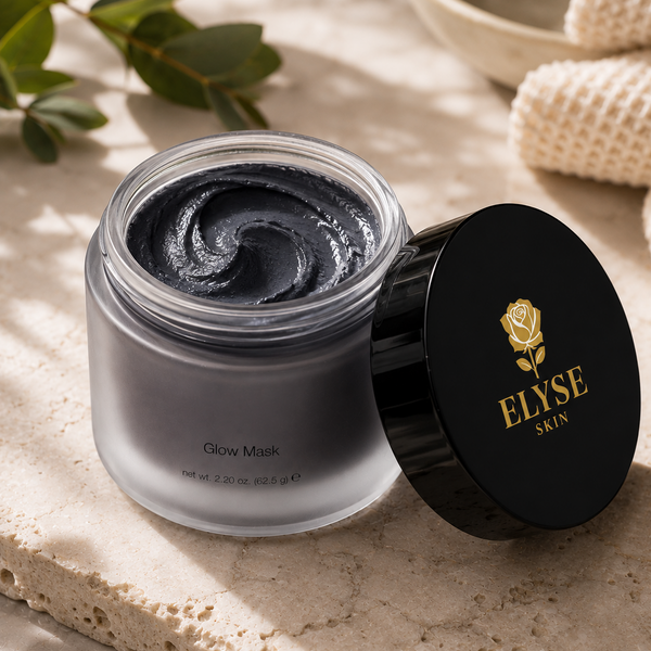 An open jar of ELYSE SKIN Glow Mask showing a dark charcoal-black mask, with its black lid resting beside it on a textured beige stone surface.