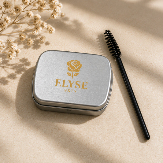 A silver metal cosmetic tin with a gold rose logo and the text 'ELYSE SKIN' sits on a beige surface next to a black spoolie brush. Dried flowers and soft shadows decorate the scene.
