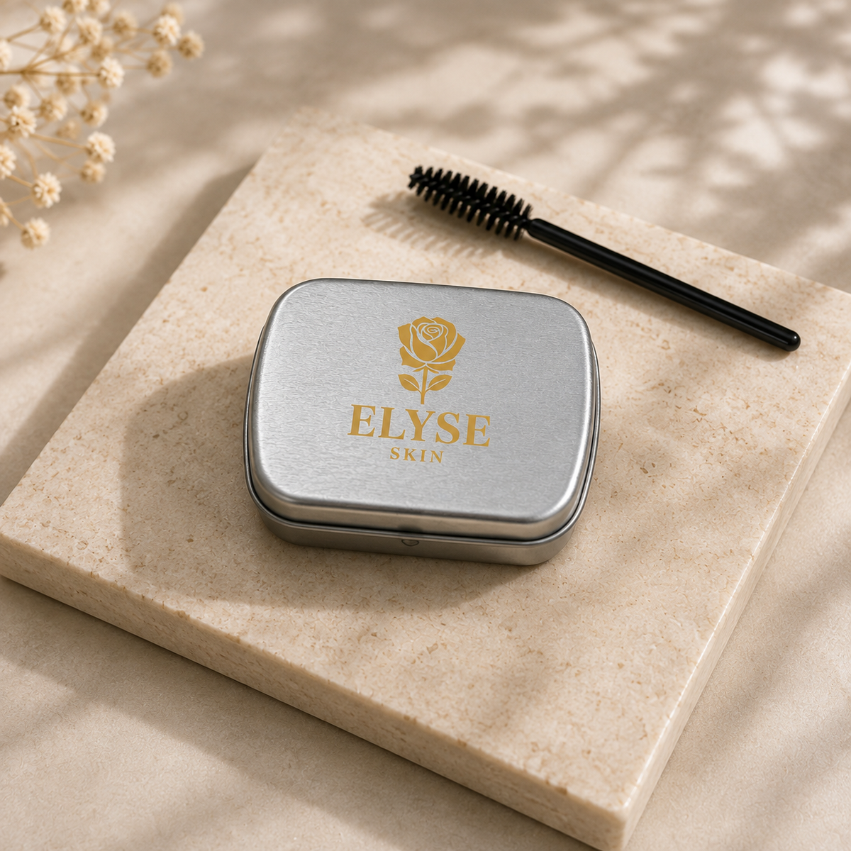 A silver metal cosmetic case with a gold rose logo and the text 'ELYSE SKIN' sits on a beige stone slab, with a black spoolie brush resting beside it and dried flowers in the corner.
