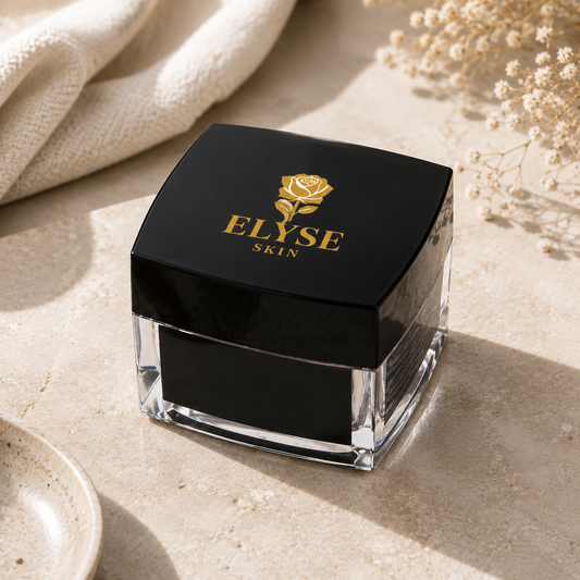 A black cosmetic jar with a clear glass base and lid, featuring the gold 'ELYSE SKIN' logo with a rose emblem, set on a beige stone surface with a cream fabric and dried flowers in the background.