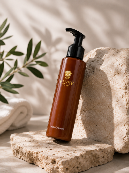 An amber brown pump bottle of ELYSE PARIS Kie` Cleanser stands on a textured stone slab with a rolled towel and green leaves in the background, under warm natural-looking shadows.