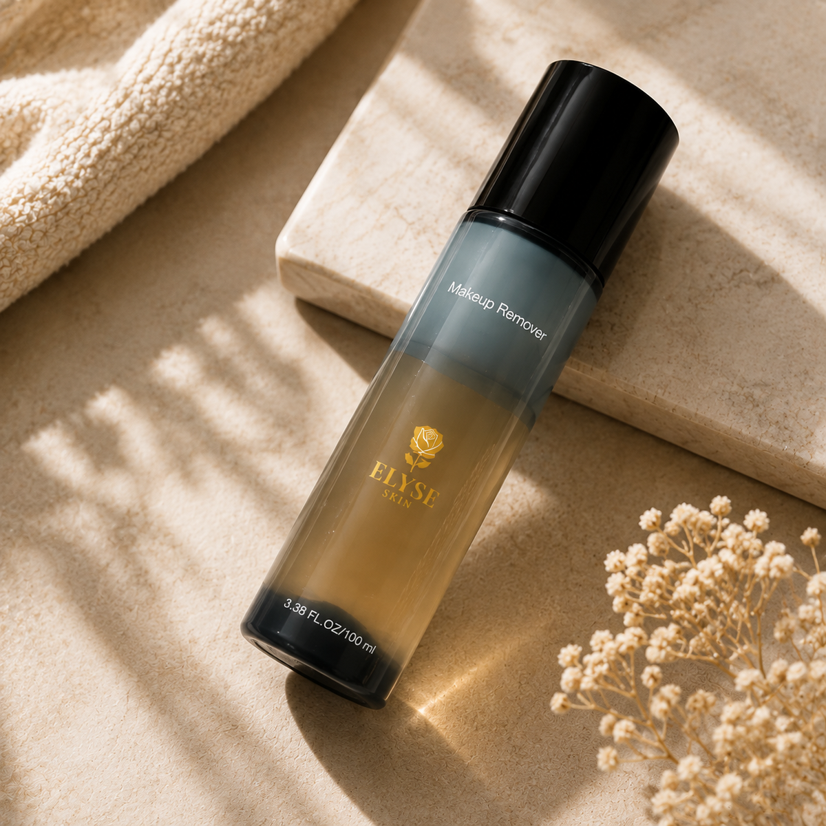 A bottle of ELYSE PARIS makeup remover with a black cap and a gradient bottle showing gold and gray tones, styled on a beige stone surface with a textured towel and dried flowers.