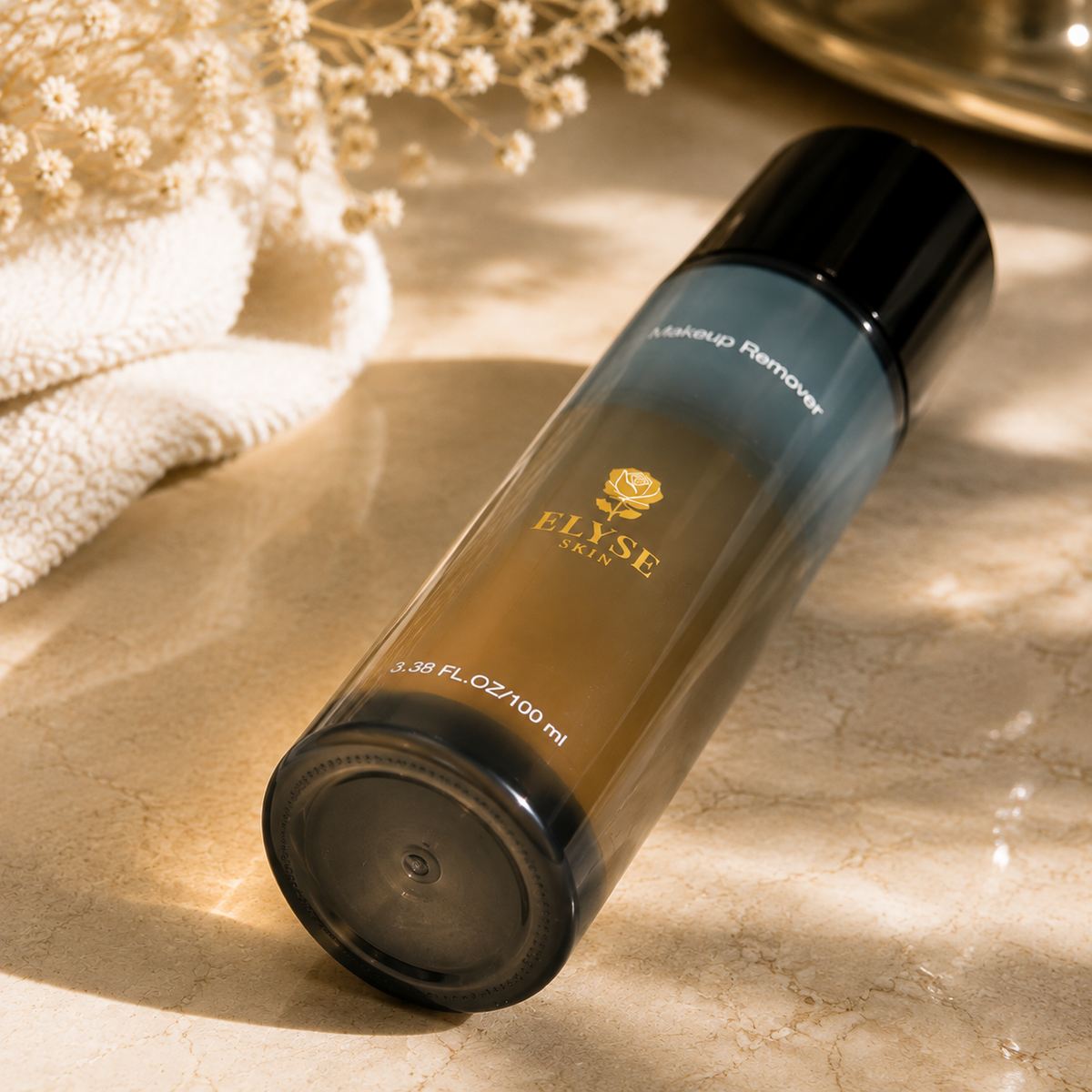 A bottle of ELYSE PARIS eyelash remover with a black cap and a gradient amber-to-dark-blue bottle, resting on a beige marble surface with dried flowers and a white towel in the background.