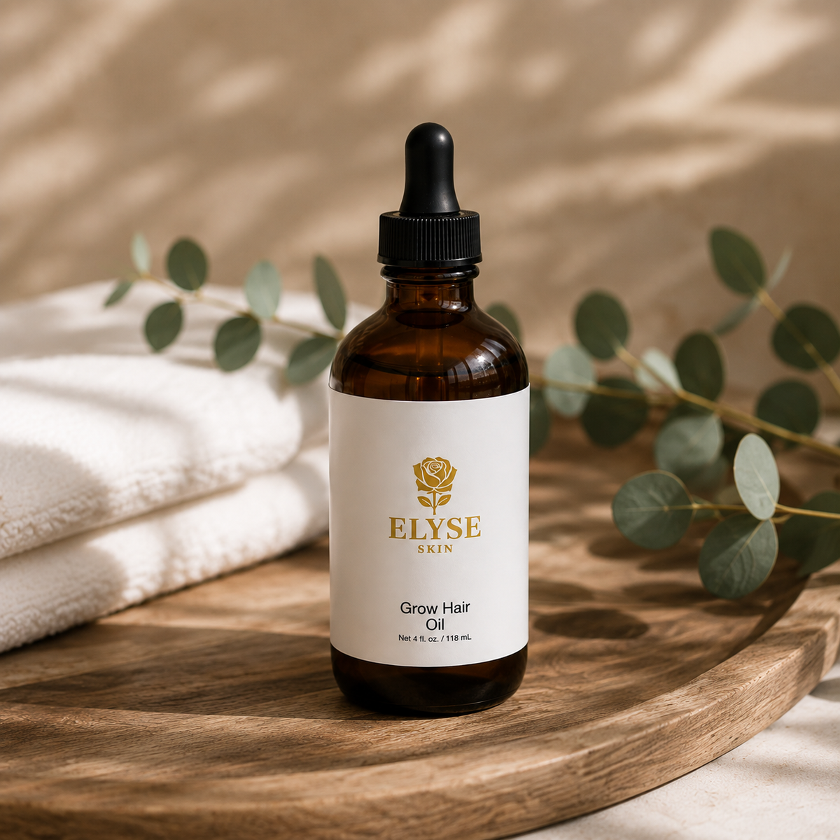An amber glass dropper bottle of ELYSE SKIN Grow Hair Oil sits on a round wooden tray, styled with a folded white towel and eucalyptus branches in soft natural light.