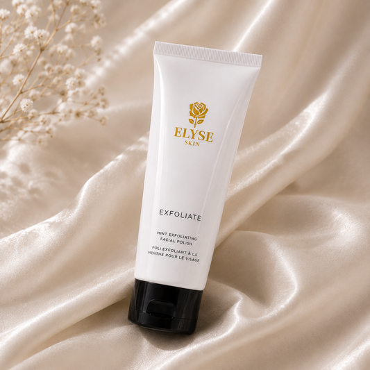 A white squeeze tube of ELYSE SKIN Exfoliate Facial Polish with a black cap, standing upright on draped champagne satin fabric with small dried flowers in the corner.
