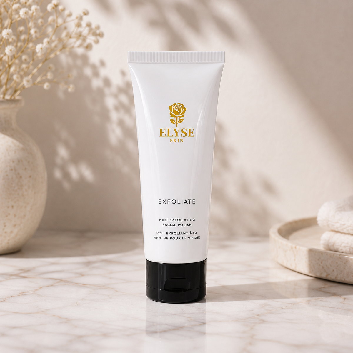 A white tube of ELYSE SKIN Exfoliate facial polish with a black cap, standing on a marble surface with soft shadows, a vase with dried flowers, and folded towels in the background.