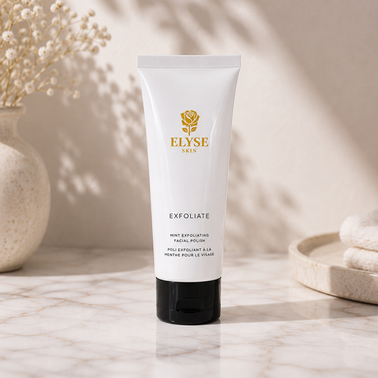 A white tube of ELYSE SKIN Exfoliate facial polish with a black cap, standing on a marble surface with soft shadows, a vase with dried flowers, and folded towels in the background.