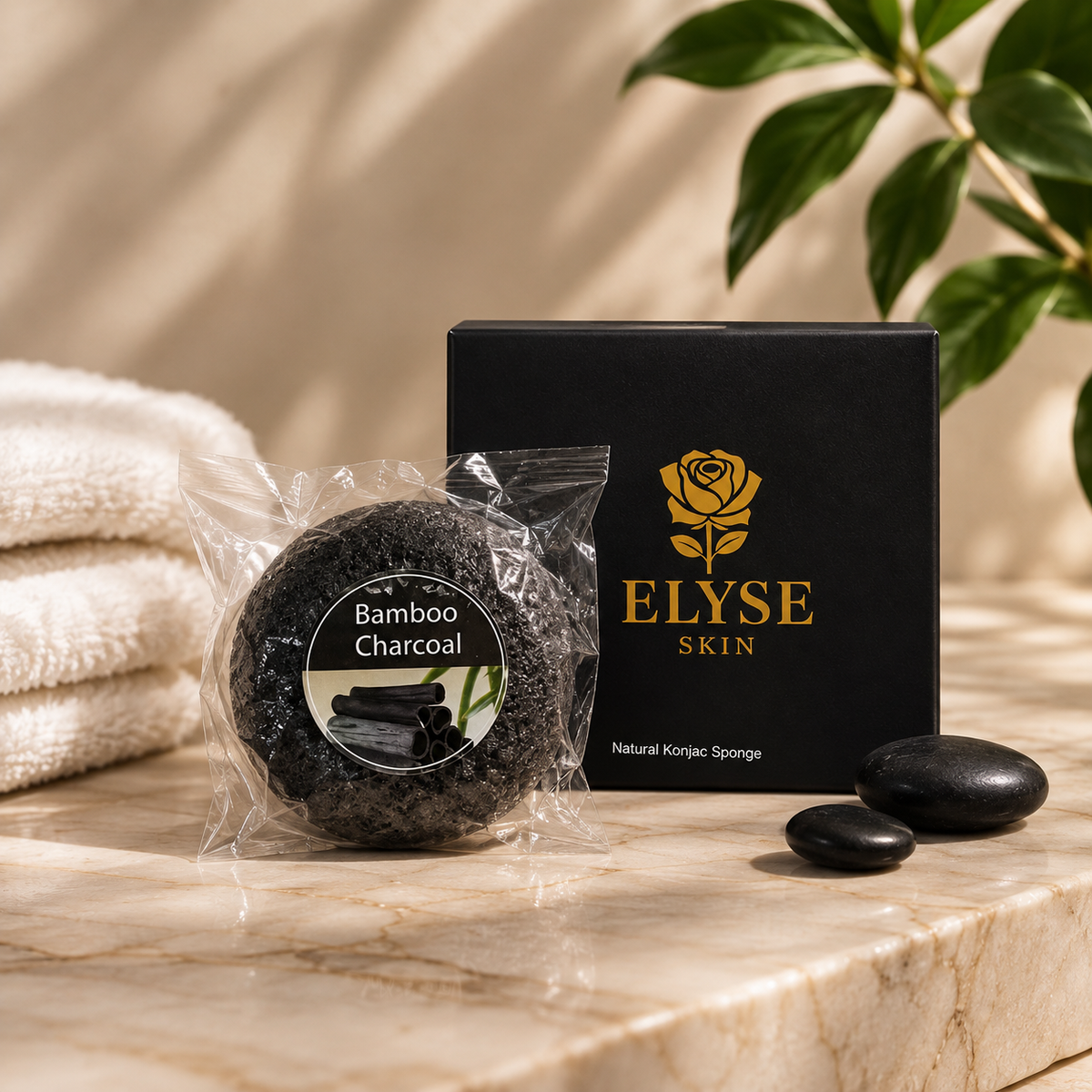 A round black bamboo charcoal sponge in a clear plastic sleeve sits in front of a black ELYSE SKIN box on a beige marble surface, with folded white towels, smooth black stones, and green leaves in the background.