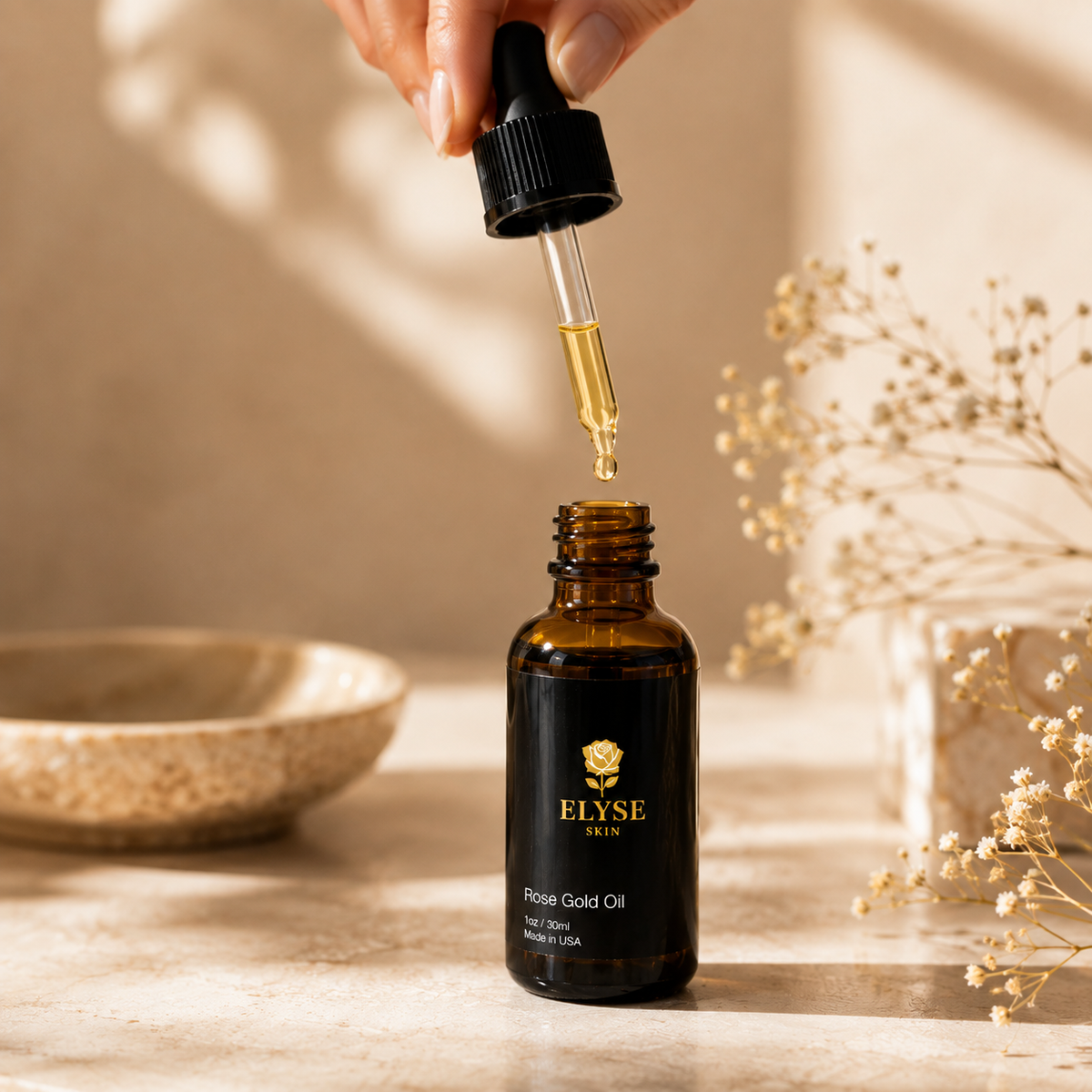 A hand holds a black dropper above an amber glass bottle labeled ELYSE SKIN Rose Gold Oil, with a gold droplet hanging over the opening. The bottle sits on a warm beige surface with dried flowers and a ceramic bowl in the background.