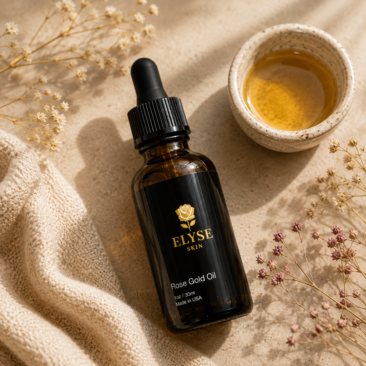An amber glass dropper bottle labeled ELYSE SKIN Rose Gold Oil, styled on a beige textured surface with dried flowers, a beige knit cloth, and a small cup of golden liquid.